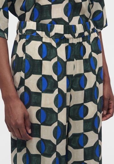 Gros plan d'une personne portant un pantalon taille haute avec une ceinture élastique et un motif géométrique dans des couleurs crème, vert foncé et bleu.