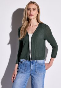 Mörkgrön, lätt cardigan med V-ringning och trekvartslånga ärmar, i kombination med ljusblå jeans som har framfickor och blekta detaljer.