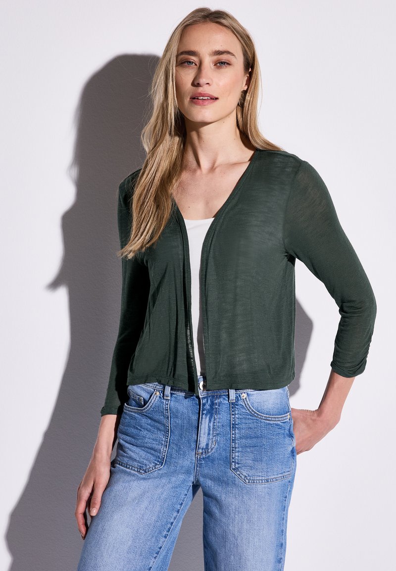 Mörkgrön, lätt cardigan med V-ringning och trekvartslånga ärmar, i kombination med ljusblå jeans som har framfickor och blekta detaljer.