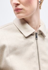 Prim-plan cu o persoană care poartă o jachetă bej cu fermoar și guler, și o cercei ovali argintii, privind într-o parte.