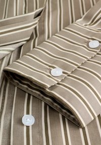 Braun- und beige gestreifter Stoff mit einer glatten Textur, der weiße Knöpfe am Ärmel aufweist. Die Streifen variieren in der Breite.