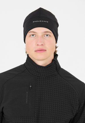 Junger Mann, der einen schwarzen Hut und eine schwarze strukturierte Sportjacke mit hohem Kragen trägt, beide mit der Aufschrift "ENDURANCE."