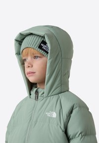 Lysegrøn dunjakke med stor hætte, lynlåslukning og logo på venstre bryst. Bæres med en matchende ribstrikket hue.