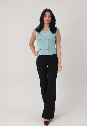 Jeune femme aux longs cheveux noirs portant un haut sans manches bleu clair à double boutonnage, un pantalon évasé noir et des chaussures pointues bordeaux, debout.