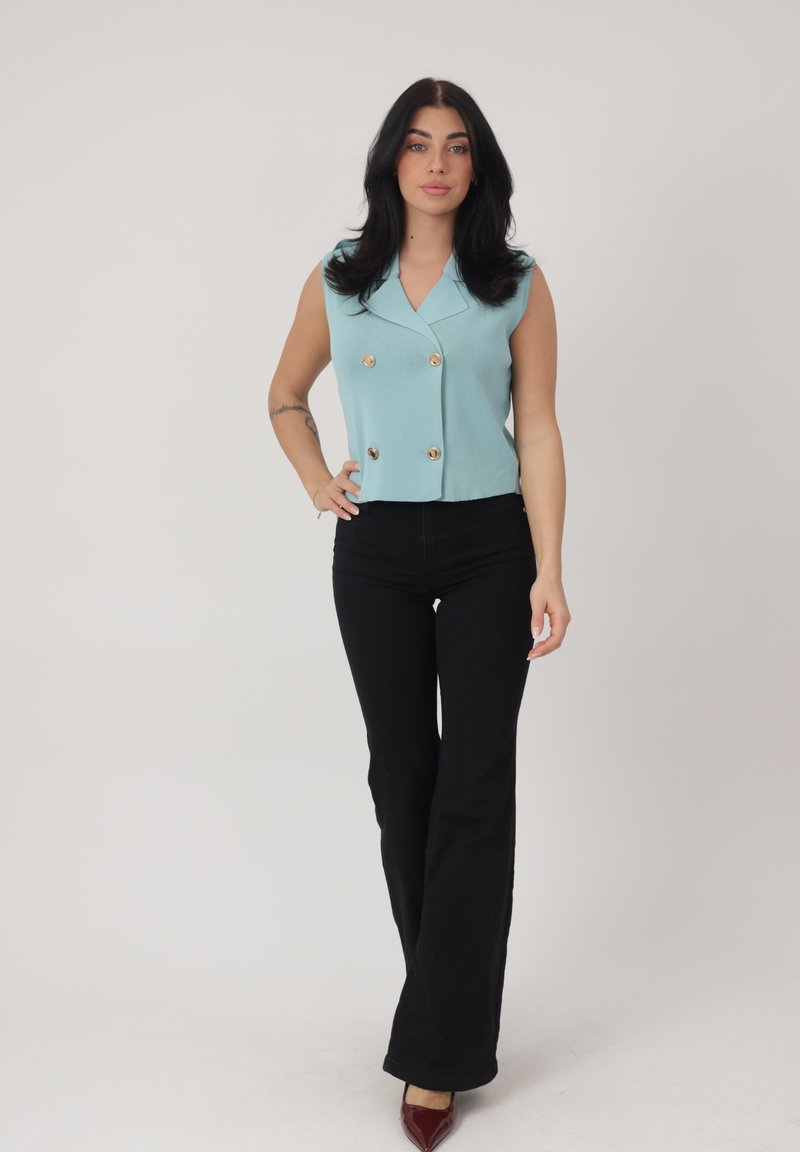 Junge Frau mit langen schwarzen Haaren trägt ein ärmelloses, hellblaues, doppelt geknöpftes Oberteil, schwarze ausgestellte Hosen und burgunderrote spitze Schuhe, steht.