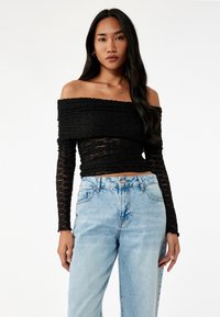 Sort off-shoulder top med struktureret stof og lange ærmer, parret med højtsiddende lyseblå jeans med knaplukning.