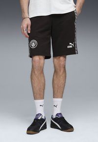 Svarta bomullsshorts med vitt Manchester City-logotyp och texten "KING". Sidostreifen har geometriska mönster. Bärs med svarta sneakers och vita strumpor.