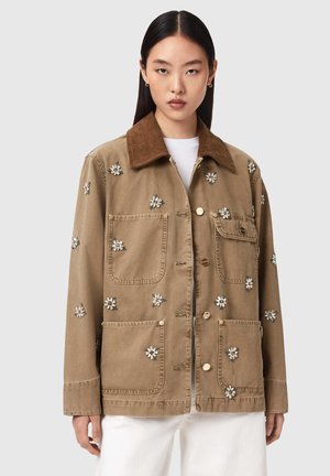 Young woman with long dark hair wears a brown jacket with large pockets and small white floral embellishments, paired with white pants.