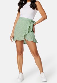 Světlě zelená květinová sukna s volánkem na spodním okraji a bočním vázáním, spárovaná s bílým žebrovaným tílkem a bílými teniskami.