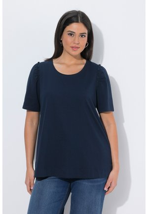 Frau mit dunklem Haar, die ein schlichtes marineblaues kurzärmeliges Oberteil und blaue Jeans trägt, vor weißem Hintergrund, frontal blickend.