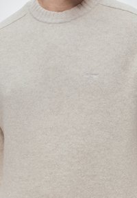 Pull beige en maille à col rond avec le logo Barbour brodé sur la poitrine, porté par une personne.