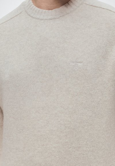 Pull beige en maille à col rond avec le logo Barbour brodé sur la poitrine, porté par une personne.