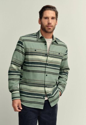 Man wearing green and beige striped button-up shirt with chest pockets, white undershirt, and dark pants, standing against a plain background.