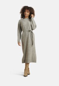 Donna con capelli ricci che indossa un lungo vestito di maglia grigio con cintura in vita e stivali al ginocchio in camoscio beige, su sfondo neutro.