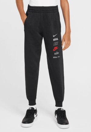 Person iført sorte Nike træningsbukser med flere Nike-logoer på højre ben, hvide sokker og sorte Nike-sneakers. Hænder i lommerne.