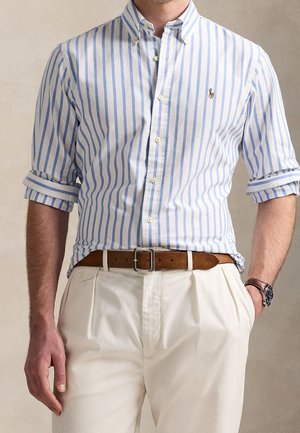 Homme portant une chemise rayée verticale bleue et blanche avec manches retroussées, pantalon plissé beige, ceinture en cuir marron et montre-bracelet.