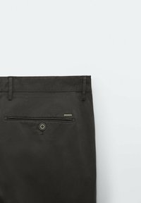 Pantalon noir avec une poche arrière fermée par un bouton et une petite étiquette métallique. Le tissu a une texture lisse et un design épuré.