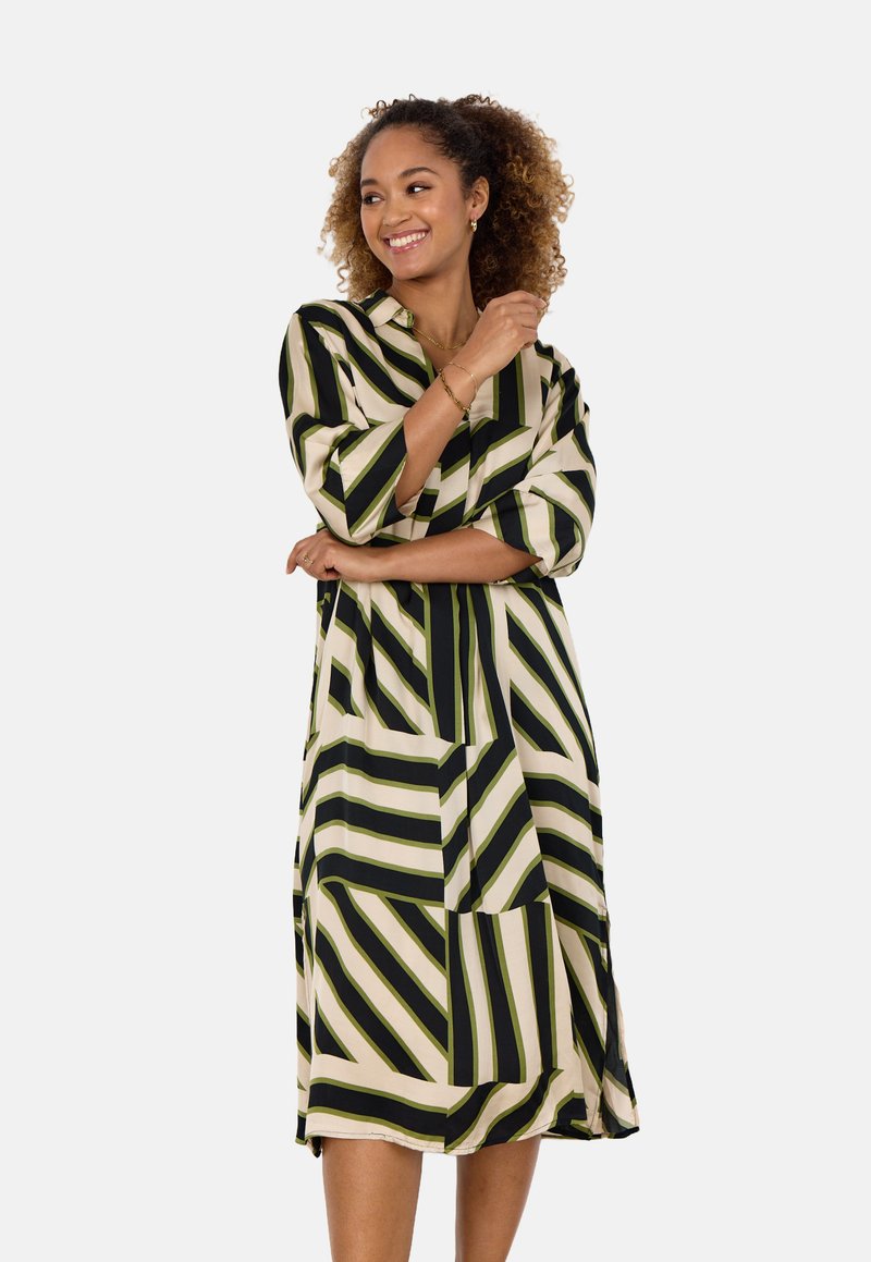 Gestreiftes Kleid mit vertikalen und diagonalen schwarzen und grünen Linien auf hellem Hintergrund, langen Ärmeln und einem lockeren Schnitt.