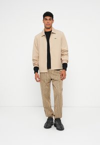 Giovane uomo in piedi, indossa una giacca beige, una camicia nera, pantaloni beige con un motivo e scarpe sportive nere, su sfondo bianco.