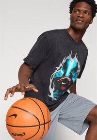 Čierne grafické tričko s modrým a plameňovým dizajnom, v kombinácii so sivými šortkami. Subjekt dribluje s oranžovou basketbalovou loptou s logom Nike.