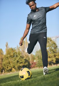 Zwarte sportbroek en een grijs shirt met korte mouwen met drie witte strepen. Een speler schopt een gele voetbal op groen gras.