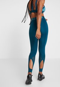 Teal urheiluliivi, jossa on leikkaus takana ja säädettävät olkaimet, yhdistettynä teal-värisiin korkea vyötäröisiin legginsseihin, joilla on verkkokankainen korostuslohko alhaalla.