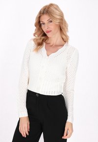 Cardigan alb tricotat, cu decolteu în V, cu nasturi în față și un model de cusătură deschis. Asortat cu pantaloni negri, subliniind designul texturat.