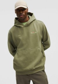 Haut à capuche vert olive avec une poche kangourou, un capuchon à cordon de serrage et un texte brodé. Fabriqué en tissu doux, porté par-dessus une chemise vert clair.