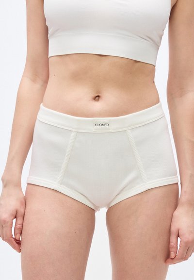 Femme portant un sous-vêtement blanc côtelé taille haute et un débardeur court avec l'étiquette "CLOSED" sur la ceinture, debout devant un fond clair.