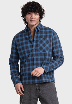 Blau- und schwarz karierte Hemd mit langen Ärmeln, Knopfkrawatte und einer Brusttasche. Aus strukturiertem Stoff gefertigt. Getragen mit grauen Jeans.