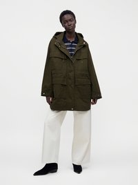Personne portant une veste à capuche ample vert foncé, une chemise rayée bleu marine, un pantalon large blanc et des bottines noires, debout devant un fond uni.