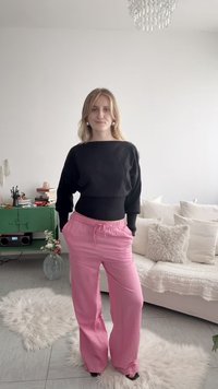 Femme debout dans un salon portant un pull noir et un pantalon rose, avec un canapé blanc, un meuble vert et des tapis gris clair visibles.
