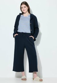 Navyblauwe hoodie met knoopaccenten over een gestreept blauw-wit shirt. Gecombineerd met navy brede broek en beige slipper.