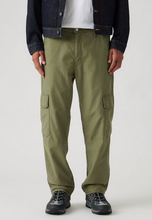 Pantalons cargo vert olive avec deux poches latérales, fermeture à bouton et coupe décontractée ; associés à des chaussures de randonnée noires et une veste en denim.