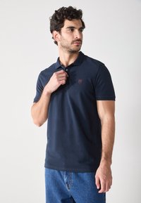 Polo shirt azul marino de algodón, con un tapeta de dos botones y un pequeño logo bordado en el pecho. Llevado con jeans de mezclilla.