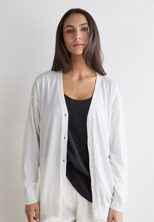 Frau mit langen braunen Haaren, die einen offenen weißen Cardigan über einem schwarzen Top und weißen Hosen trägt, steht vor einer schlichten hellen Wand.