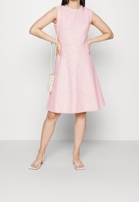 Vaaleanpunainen olkaimeton mekko, jossa on teksturoitu kangas, halkiotettu hame ja frillattuja olkaimia. Yhdistetty pieneen vaaleaan laukkuun ja remmisandaaleihin.