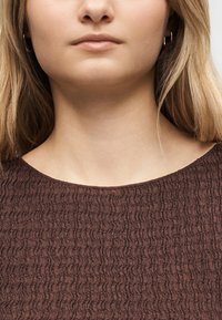 Κοντινό πλάνο του κάτω μέρους του προσώπου και του λαιμού ενός ατόμου με ανοιχτόχρωμο δέρμα, που φοράει καφέ υφασμάτινο τοπ και μικρά ασημένια κρίκους σκουλαρίκια.