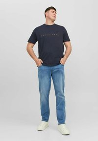 Navyblauwe katoenen t-shirt met het "JACK & JONES" logo in oranje, gecombineerd met lichtblauwe jeans en witte sneakers, staande pose tegen een effen achtergrond.