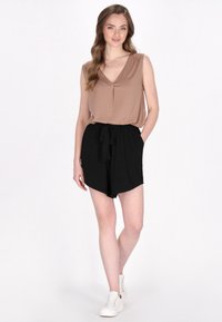 Top marrone senza maniche con scollatura a V, abbinato a pantaloni corti neri a vita alta con cintura annodata. Il modello indossa sneakers bianche.