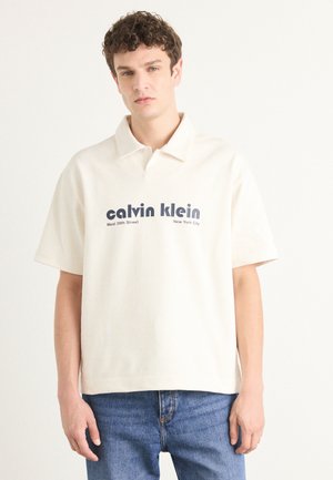 Beige polo shirt with a collar, featuring a graphic print in navy blue saying "calvin klein" and "West 39th Street, New York City."