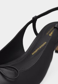 Μαύρο δερμάτινο παπούτσι slingback με μυτερή διακοσμητική φιόγκο στη μύτη και χαμηλό χοντρό τακούνι. Ορατό χρυσό κείμενο "Contour Cushion" στο εσωτερικό.