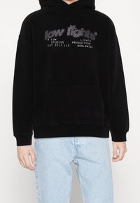 Hoodie noir en tissu doux avec une poche avant, doté d'un logo texte réfléchissant en argent. Poignets et ourlet côtelés complètent le design.