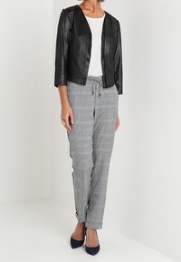 Femme portant une veste courte noire, un haut blanc, un pantalon gris à carreaux avec cordon de serrage, et des escarpins bleu foncé à bout pointu, posant devant un fond blanc.