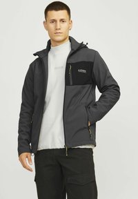 Zwarte softshelljack met een ritsborstzak, voorzakken, elastische manchetten en een verstelbare zoom, gecombineerd met een lichtgrijze sweatshirt.