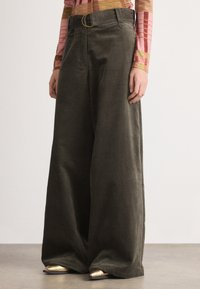 Pantalons larges en velours côtelé vert olive, avec une taille haute, des passants de ceinture et une grande boucle de ceinture ronde. Texture lisse et coupe ajustée.