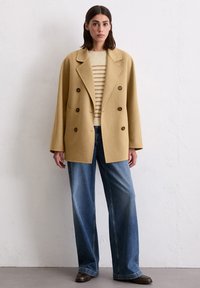 Double-breasted beige coat with large buttons, worn over a cream striped sweater and wide-leg blue jeans. Smooth texture, relaxed fit.