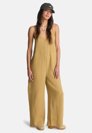 Musztardowożółty kombinezon z lekkiego, teksturowanego materiału, szerokie nogawki i głęboki dekolt w kształcie litery V. Połączony z ciemną czapką i neutralnymi chodakami.