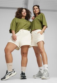 Dva modela nose oversized maslinasto zelene majice i krem kratke hlače. Imaju usklađene tenisice i lagane čarape. Jednostavna, ležerna atletika.
