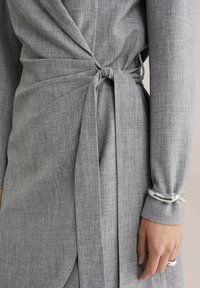 Hall halli rüütelkleit tekstureeritud kangaga, millel on vöökohal lips ja pikad varrukad, täiustatud hõbedase käevõru abil randmel.